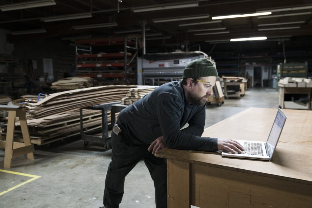 A Caucasian carpenter working on his lap top after hours in a large woodworking factory.